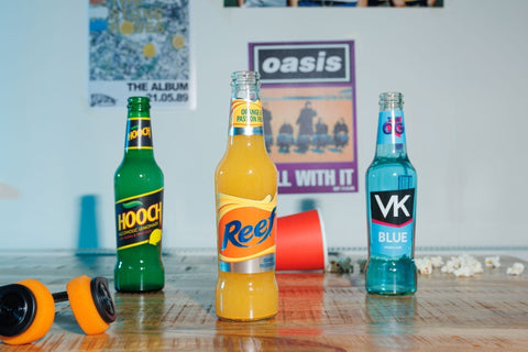Bottle of Lemon Hooch, Reef Orange & Passionfruit and VK Blue on a table in front of 90s posters including Oasis Roll With It, The Stone Roses...