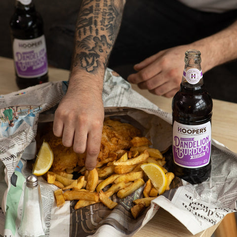 Fish and chips wrapped traditionally in a newspaper with Hoopers Alcoholic Dandelion & Burdock drink, featuring golden chips, fresh lemon wedges on a wooden table.