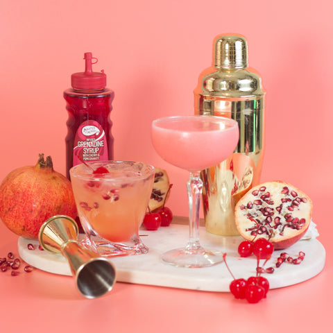 Cocktails made with Master of mixes cocktail essentials blue curacao, perfect for making cocktails and mocktails. Surrounded by pomegranates, cherries and a cocktail shaker. 