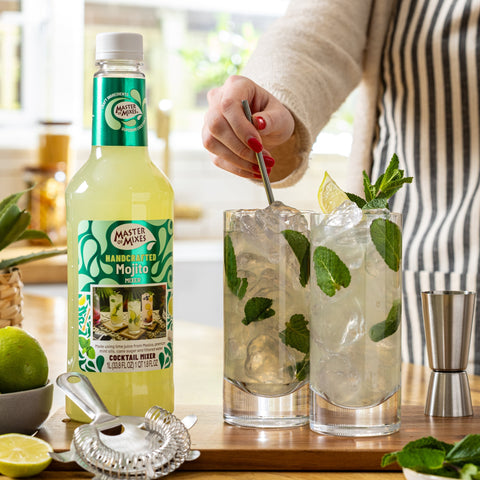 Person preparing a mojito cocktail with a bottle of Master of Mixes Mojito mix on a kitchen counter.