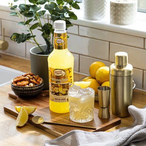 Bottle of Master of Mixes Whiskey Sour mix on a kitchen counter with a cocktail glass and lemons.