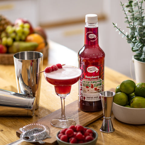 Bottle of Master of Mixes Raspberry with a cocktail glass, limes, and other bar tools on a wooden surface.
