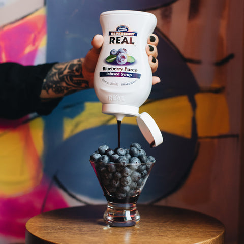 Someone pouring Real blueberry puree syrup over a bowl of blueberries, Real syrups are perfect for cooking & baking, making cocktails or mocktails and dirty sodas. 