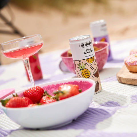 Can of All Shook Up Pina Colada with a bowl of strawberries and a pink drink in the background.