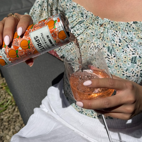A person pouring a ready to drink All Shook Up peach and lychee white wine spritz into a glass outside. 