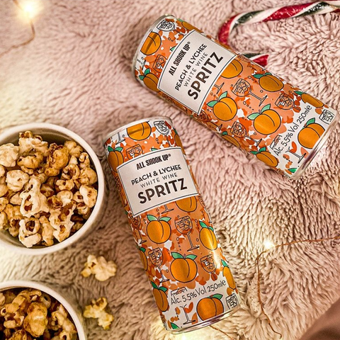 Two ready to drink cans of peach and lychee white wine spritz next to a bowl of popcorn. 