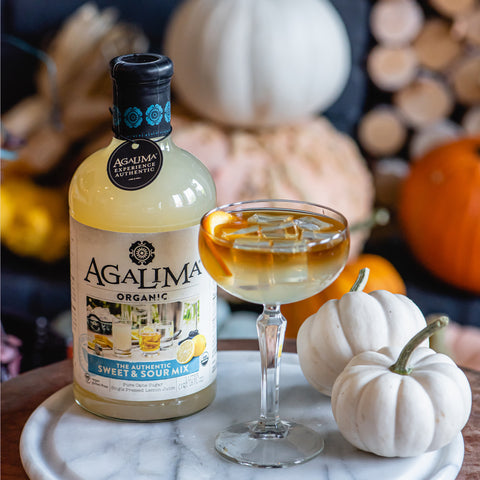 A 1L glass bottle of Agalima Organic Bloody Mary Mix next to a sweet & sour cocktail in an autumn-themed setting.