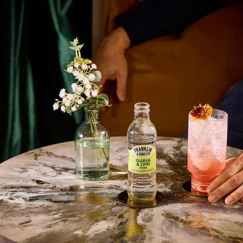 A glass bottle of Franklin & Sons guava & lime soda, a premium soft drink, next to someone drinking it in a glass as a mixer.  