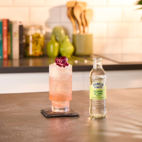 A glass bottle of Franklin & Sons guava & lime soda, a premium soft drink, next to someone drinking it in a glass as a mixer. 