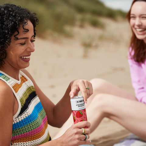Two women having a picnic on the beach with one of them opening All Shook Up strawberry daiquiri premixed, ready to drink canned cocktail. 