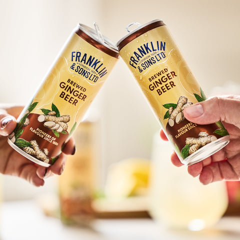 Two cans of Franklin & Sons Ginger Beer being held in a celebratory manner with a blurred background.
