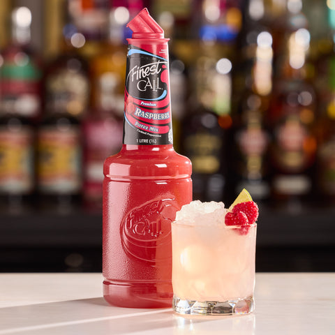 Red bottle of Finest Call Raspberry Puree next to a cocktail garnished with raspberries on a bar counter.