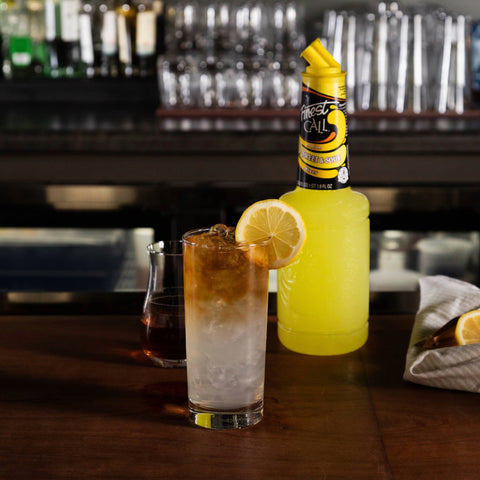 Bottle of Finest Call Sweet & Sour next to a drink in a bar background. 