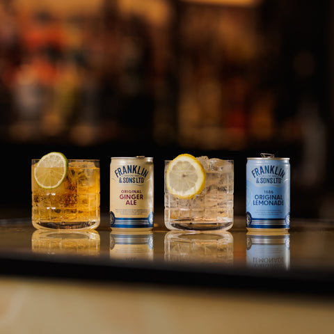 Two Franklin & Sons cans in flavours: 1886 original lemonade and original ginger ale, next to glasses filled with each of them. 