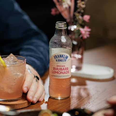 Bottle of Franklin & Sons Spring Rhubarb Lemonade on a table with a drink and flowers in the background.