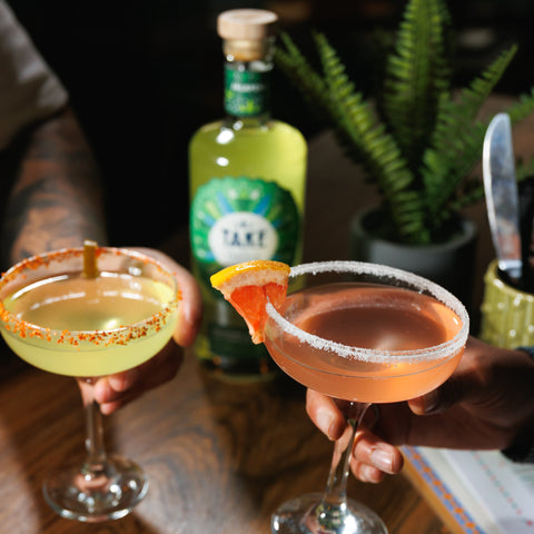 Two people drinking cocktails made with Take tequila, with the bottle of take jalapeno tequila liqueur in the background.
