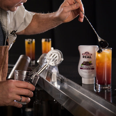 Squeezy bottle of Black Cherry Puree Re'al Infused Exotics Syrup next to a cocktail in a bar setting.