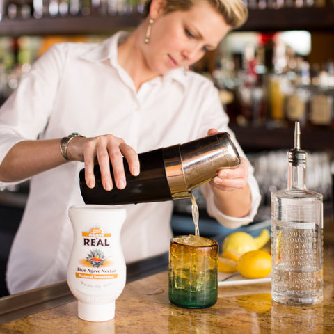 Squeezy bottle of Blue Agave Nectar Re'al Infused Exotics Syrup next to a bartender serving a cocktail in a bar setting.