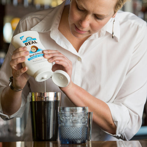 Bottle of gourmet Cream of Coconut Coco Re'al being squeezed by a bartender into a cocktail shaker.