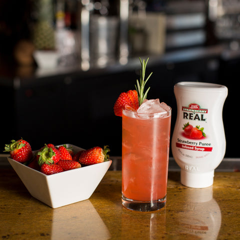 Squeezy bottle of Strawberry Puree Re'al Infused Exotics Syrup next to a strawberry themed cocktail with a owl of ripe juicy strawberries.