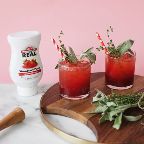 Squeezy bottle of Strawberry Puree Re'al Infused Exotics Syrup next to a wooden board with two strawberry cocktails with straws.