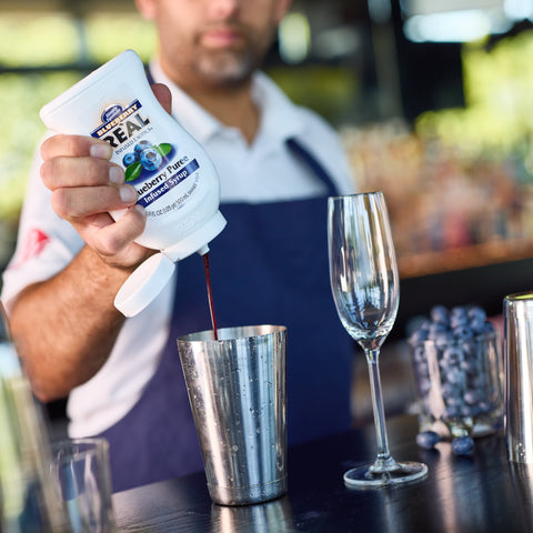 Person squeezing Real Blueberry Puree into a cocktail shaker at a bar.