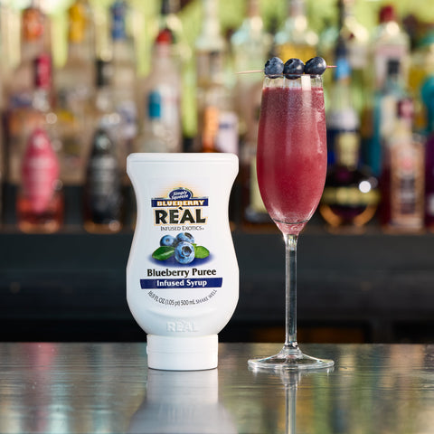 A bottle of Real Blueberry Puree with a cocktail garnished with blueberries on a bar counter.