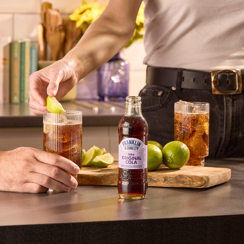 Two people drinking glasses of Franklin &  Sons 1886 original cola, with the Franklin & Sons 1886 original cola bottle in the front. 