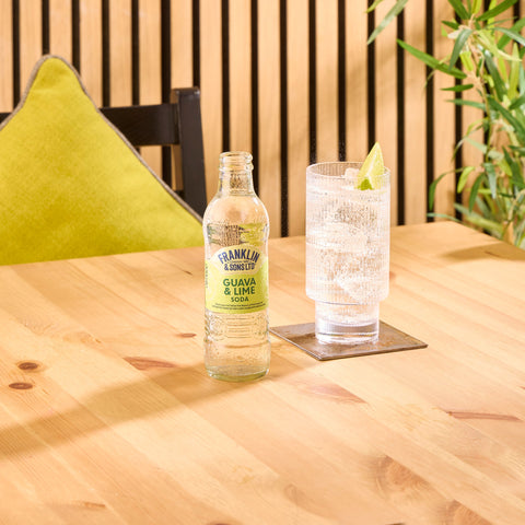 A glass bottle of Franklin & Sons guava & lime soda, a premium soft drink, next to a glass of the soda. 