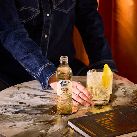 A glass bottle of Franklin & Sons original ginger ale, a premium soft drink & mixer, with someone drinking a glass of the ginger ale. 