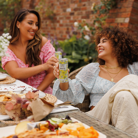 Two women sat outside at a picnic drinking All Shook Up Elderflower & Mint white wine spritz canned cocktails.