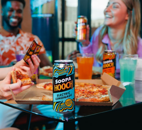 Friends enjoying pizza with cans of Soopa Hooch with the Electric Lemonade flavour being the foreground. People are laughing and playing card games during a social gathering.