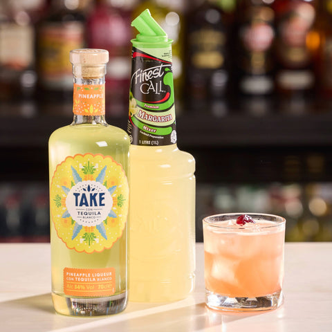 A bottle of Take Tequila Pineapple, a bottle of Finest Call Margarita Mix, and a cocktail on a bar counter.