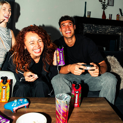 Three people sitting together, laughing and playing video games with gaming controllers and drinks on a table. With packs of VK Squashka.