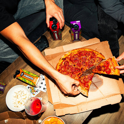 Person taking a slice of pizza from a box with drinks and snacks on a table.