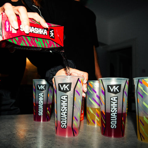 Person pouring VK Squashka drinks into clear plastic cups on a dark surface.