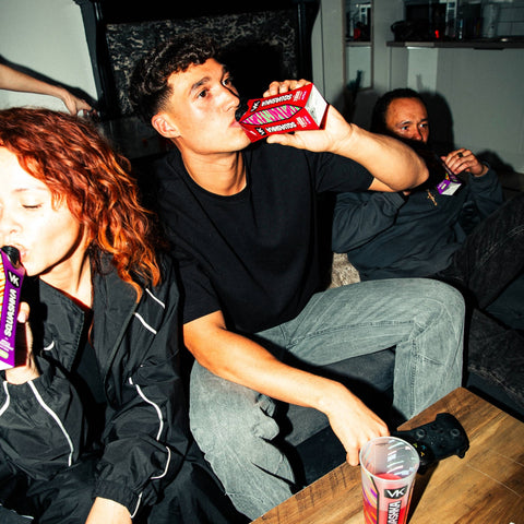 Two people sitting together, one holding a red box of VK Squashka and the other a purple carton of VK Squashka, with a camera on a table in front of them.