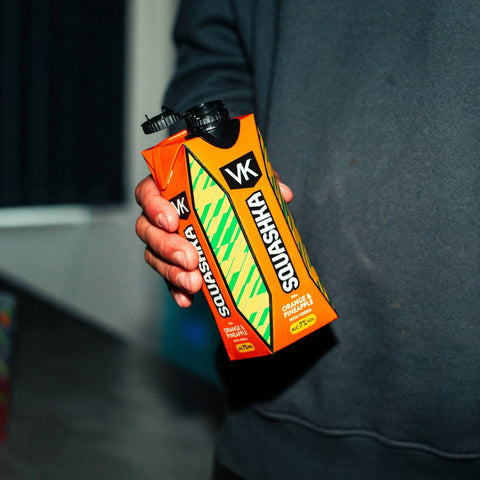 Hand holding a VK Squashka orange & Pineapple drink package against a dark background