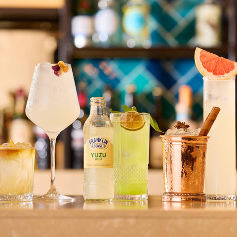 Beverages including a bottle of Franklin & Sons Yuzu on a bar counter with various cocktails.