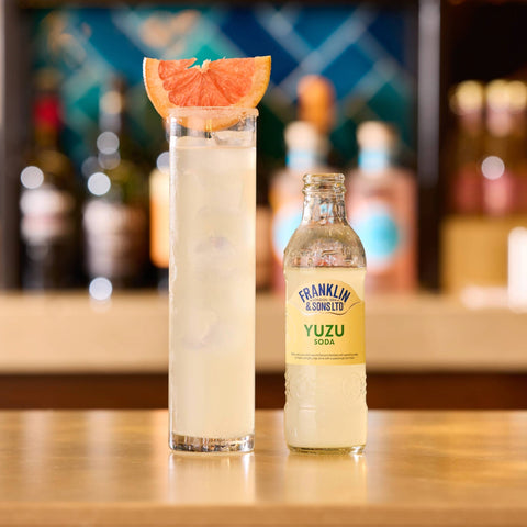 Bottle of Franklin & Sons Yuzu Soda next to a tall glass with a drink and grapefruit slice on a bar.