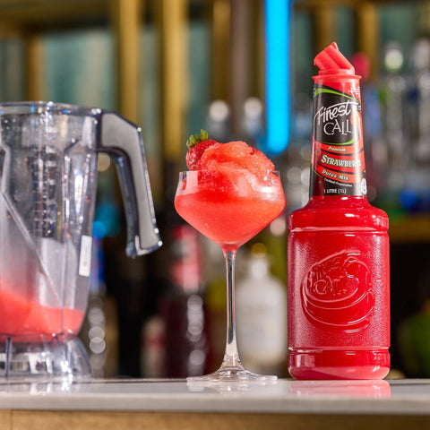 Red frozen cocktail in a glass with a bottle of Finest Call Strawberry Puree on a bar counter.