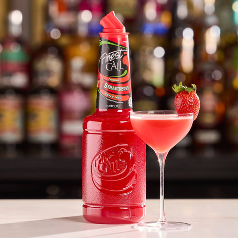 Red bottle of Finest Call strawberry puree with a strawberry garnish on a cocktail glass filled with a strawberry daiquiri on a bar background.