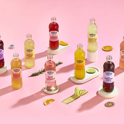 Multiple bottles of Franklin & Sons soft drinks on a pink background with different fruits around the bottles, Franklin and Sons offer premium soft drinks which are good for mixing. 