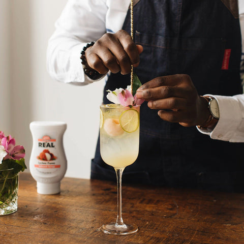 Someone making a lychee cocktail using Re'al Lychee Puree Infused Syrup, with the Re'al syrup bottle in the background. 