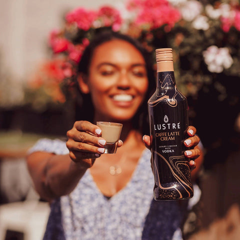 A woman holding a shot of Lustre Caffe Latte Cream Vodka Liqueur, a vegan cream liqueur, also holding the bottle of Lustre. 