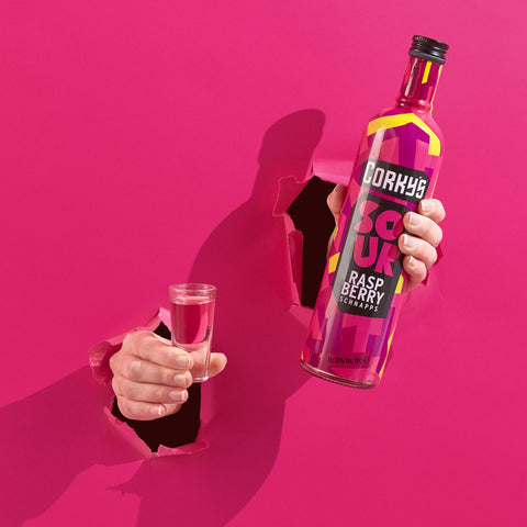 A bottle of Corky's sour raspberry schnapps and a shot of the schnapps coming through a bright pink background. Corky's is perfect for easy shots and cocktails. 