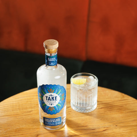 Bottle of 'Take' Tequila with a glass on a wooden table against an orange cushioned background