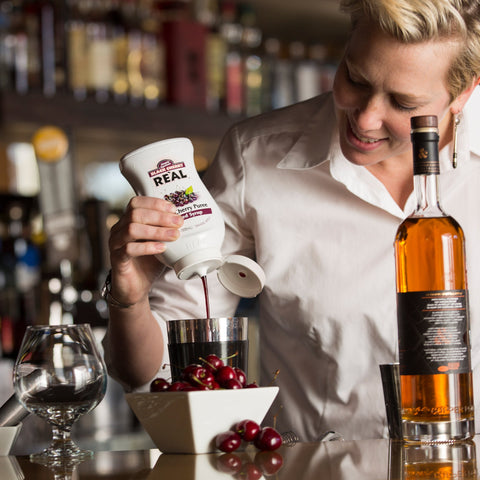 Squeezy bottle of Black Cherry Puree Re'al Infused Exotics Syrup being squeezed into a cocktail shaker.