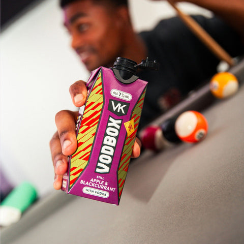 Someone holding a carton of VK VODBOX Apple & Blackcurrant, a premixed alcopop, whilst playing pool.    