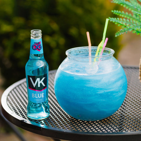 A bottle of VK blue next to a fishbowl of VK blue with different coloured straws on a table outside. 